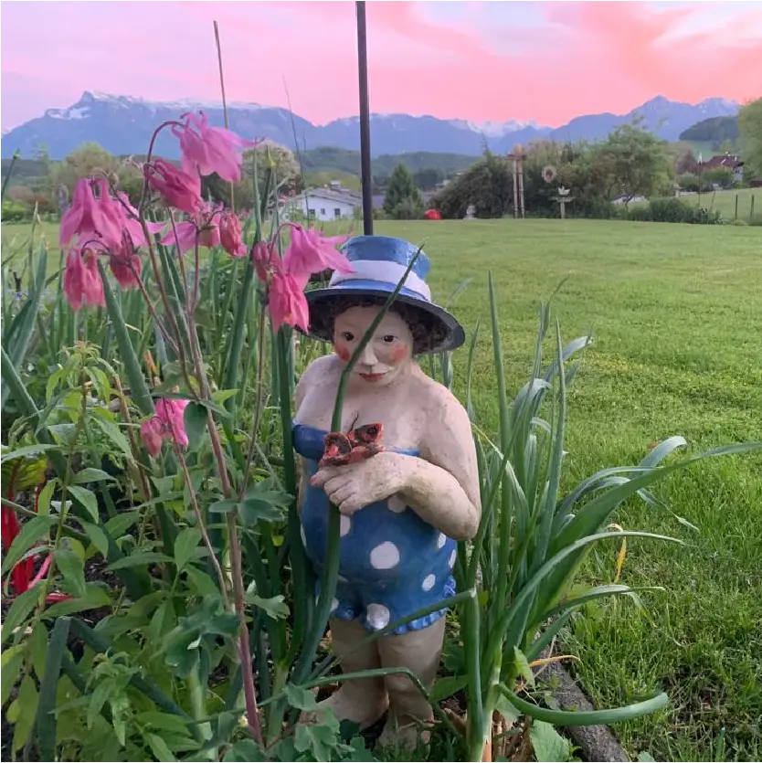 Eine Frau im Badeanzug aus Keramik in einem Blumenbeet vor einem Bergpanorama bei Sonnenuntergang.