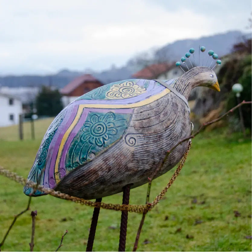 Der "Paradiesvogel": Ein Vogel aus Keramik von der Größe einer Wassermelone mit einem kurzen Hals und Kopfschmuck aus bunten Perlen auf Draht. Der Vogel steht auf zwei Metallstangen in einer Wiese.